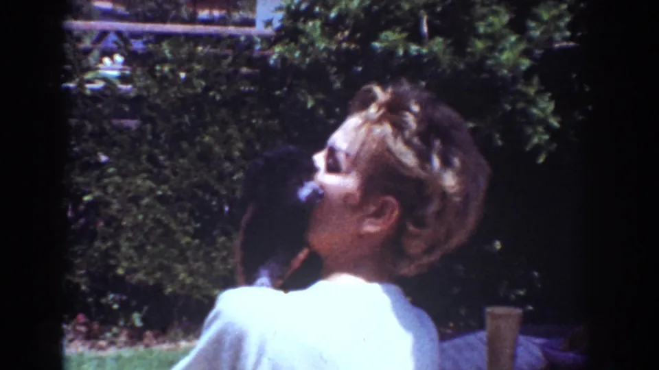 Wider 1962 Yorba Linda home-movie view of the woman holding the puppy, with lawn, foliage, and patio items visible.