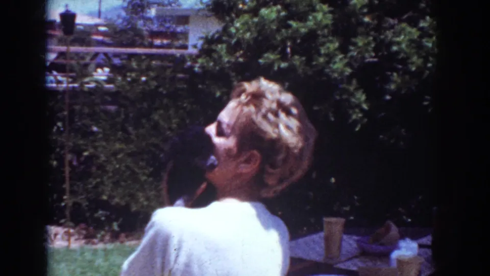1962 Yorba Linda frame showing the woman and black puppy in a backyard, with foliage, lawn, and patio objects in view.