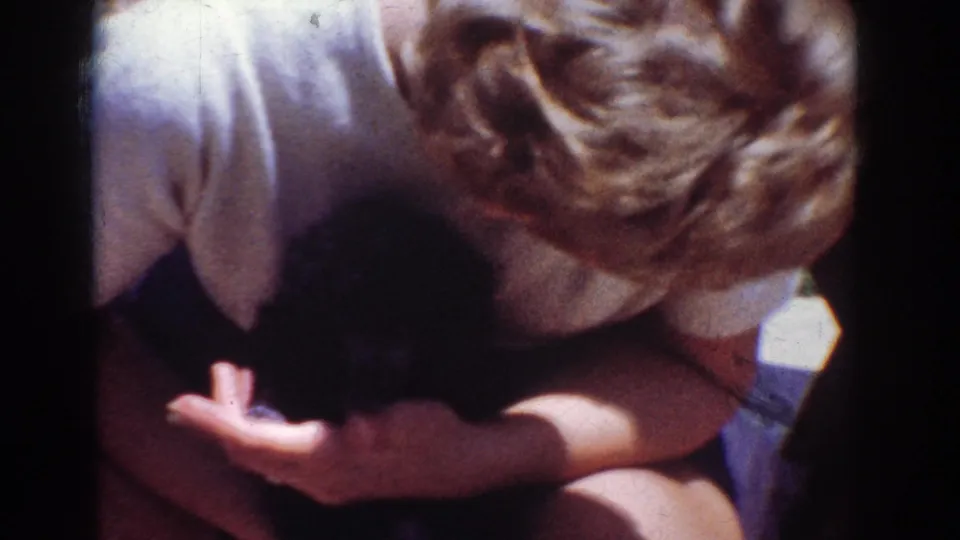 The woman holds the small black puppy close while extending one hand, likely offering a treat in the 1962 home-movie scene.