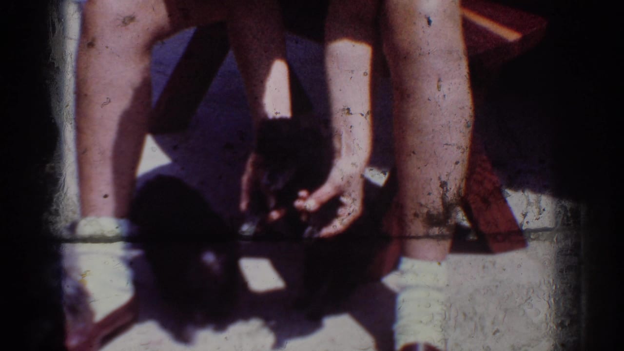 1962 Yorba Linda home-movie frame showing a person’s legs and hands near a small black puppy on a sunlit wooden deck.