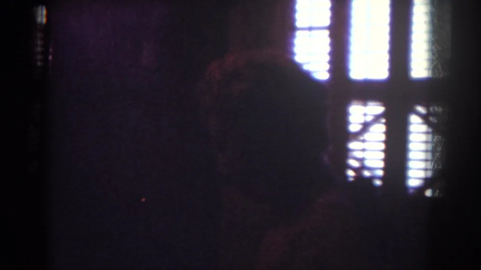 Dark 8mm interior frame with a silhouetted figure or shape near bright window blinds, likely from the same Yorba Linda home-movie sequence.