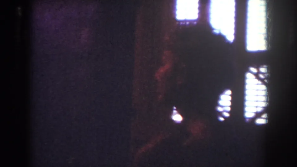 Dim home-movie interior frame with light filtering through blinds and a faint reddish silhouette against the wall.