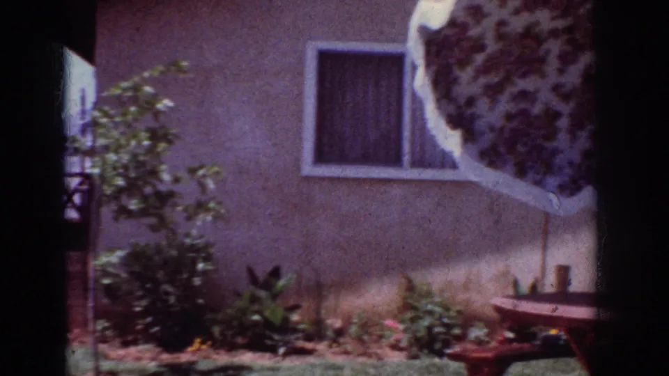 A floral patio umbrella enters the frame near a curtained window, low plantings, and a picnic table in a 1962 backyard scene.