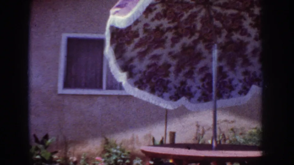 1962 Yorba Linda backyard frame showing a floral patio umbrella, picnic table, stucco wall, curtained window, and low plants.