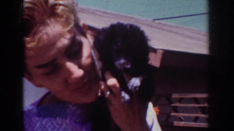 In 1962 Yorba Linda footage, an adult man holds a small black puppy close to his face outside in bright sun.