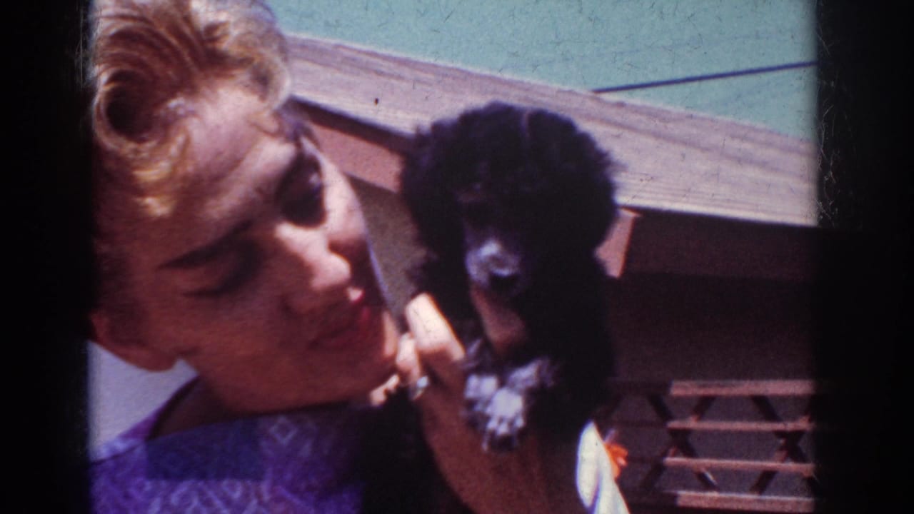 In 1962 Yorba Linda home-movie footage, a blonde woman in a purple patterned top holds a small black poodle puppy near her face outdoors.