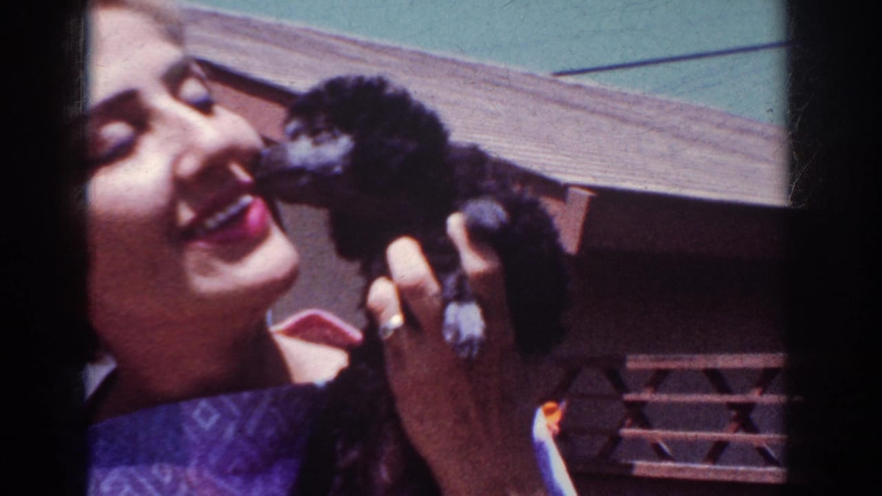 A smiling woman in Yorba Linda, California, lets a small black poodle puppy nuzzle her face in a sunlit 1962 home-movie frame.