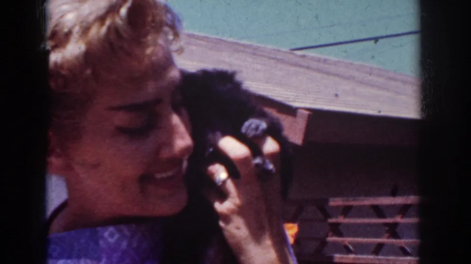 The 1962 outdoor home-movie frame shows a woman smiling as she holds a small black poodle puppy beside her cheek near a house exterior.
