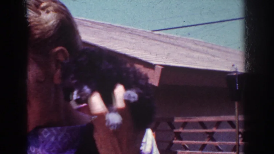 A woman turns partly away from the camera while holding a small black poodle puppy in sunlit 1962 Yorba Linda home-movie footage.
