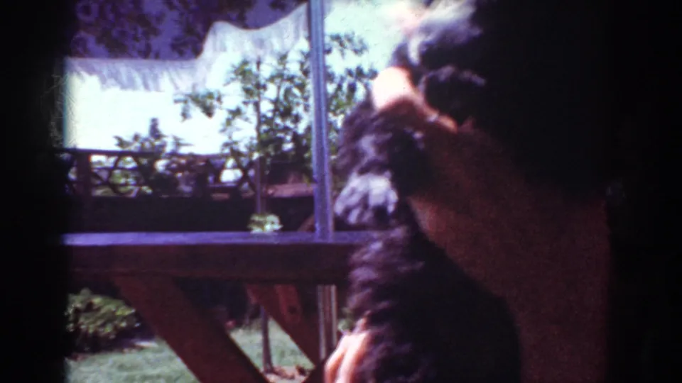 A woman holds the small black poodle close to her face on a sunny patio in 1962 Yorba Linda home-movie footage.