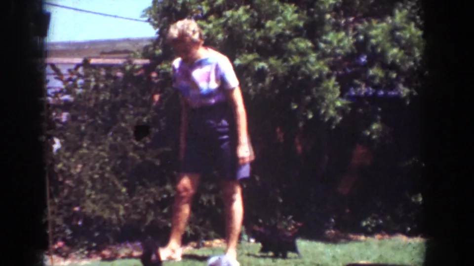 A woman in blue stands in a sunny Yorba Linda backyard as a small dark dog remains near the shrubs in this 1962 home-movie frame.