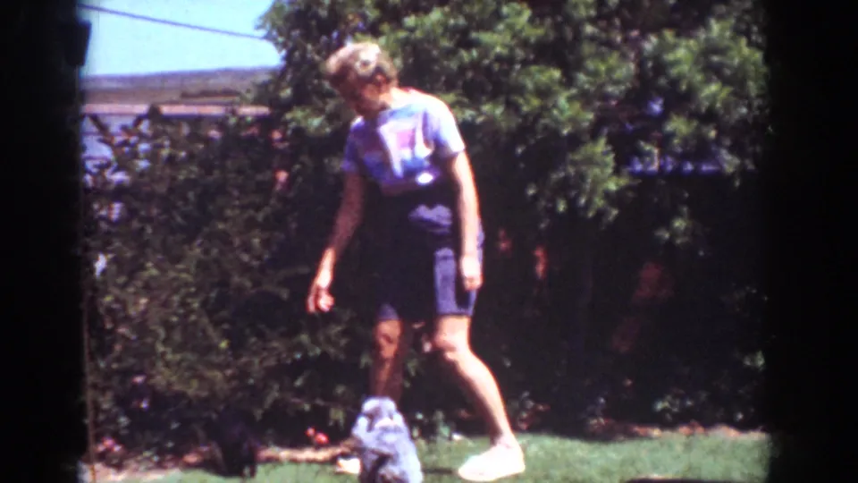 The woman moves across the Yorba Linda lawn while the small dark dog stays by the bushes, matching the supplied 1962 backyard context.