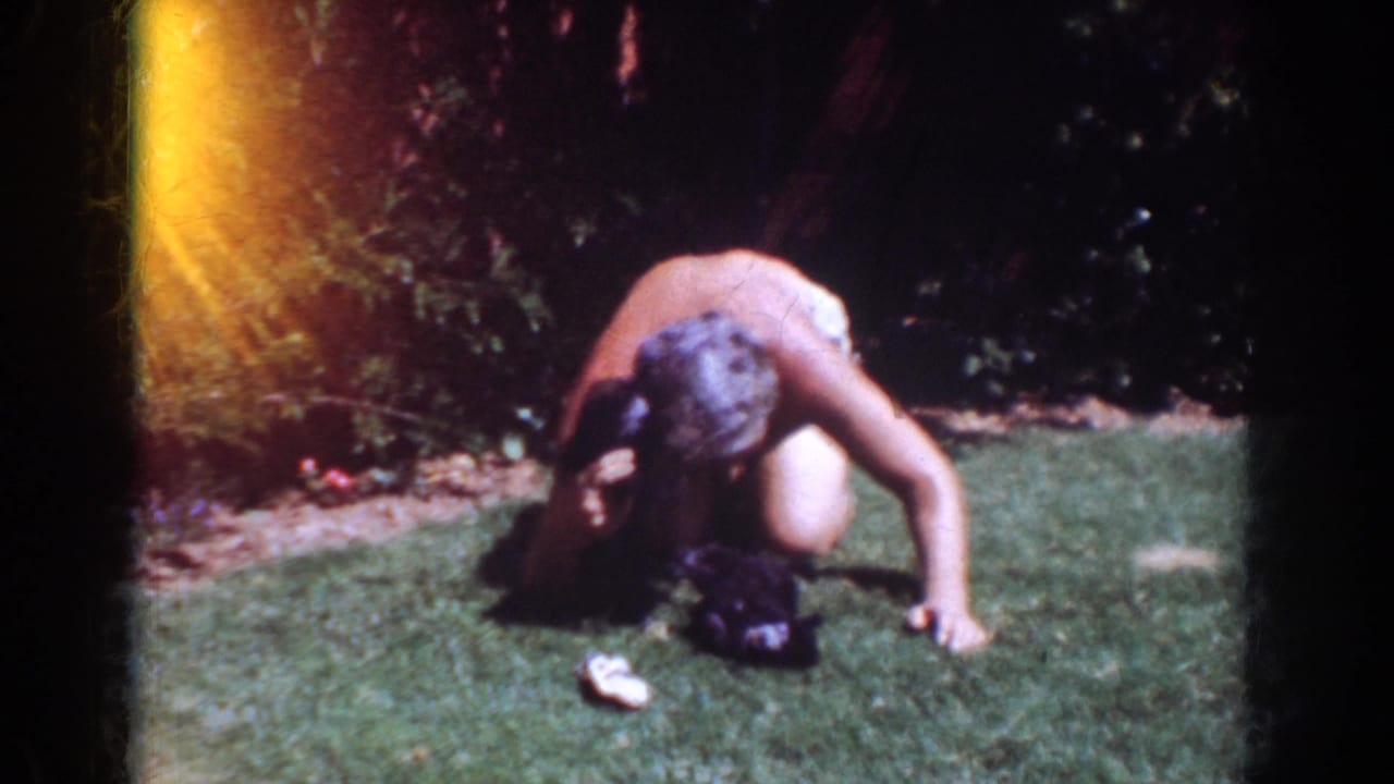 In 1962 Yorba Linda home-movie footage, an older shirtless man kneels on grass while playing with a small black puppy.