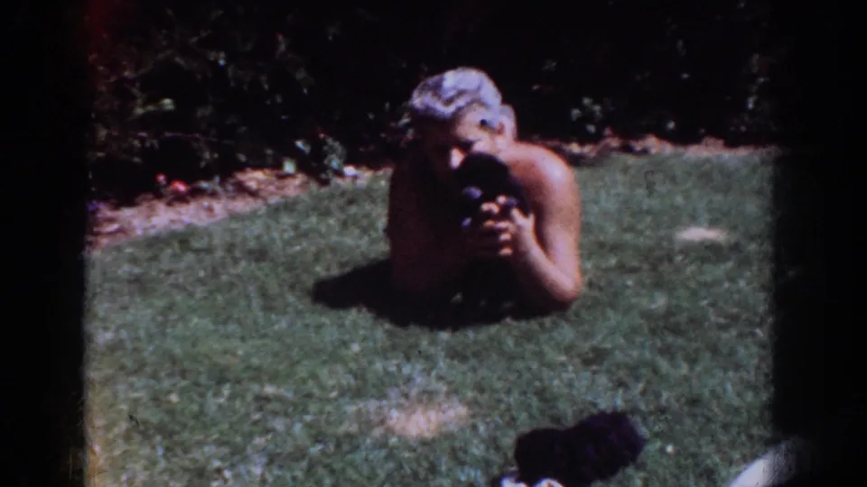 An older man lies on a Yorba Linda lawn, holding a small black puppy close in a warm 1960s home-movie frame.
