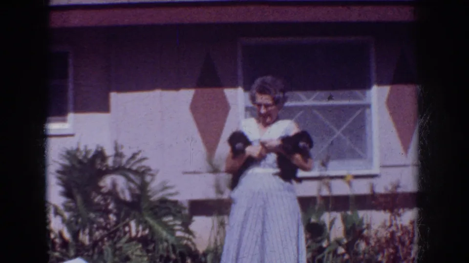 A woman in a pale dress stands before a house in Yorba Linda, holding two dark puppies in 1962 home-movie footage.