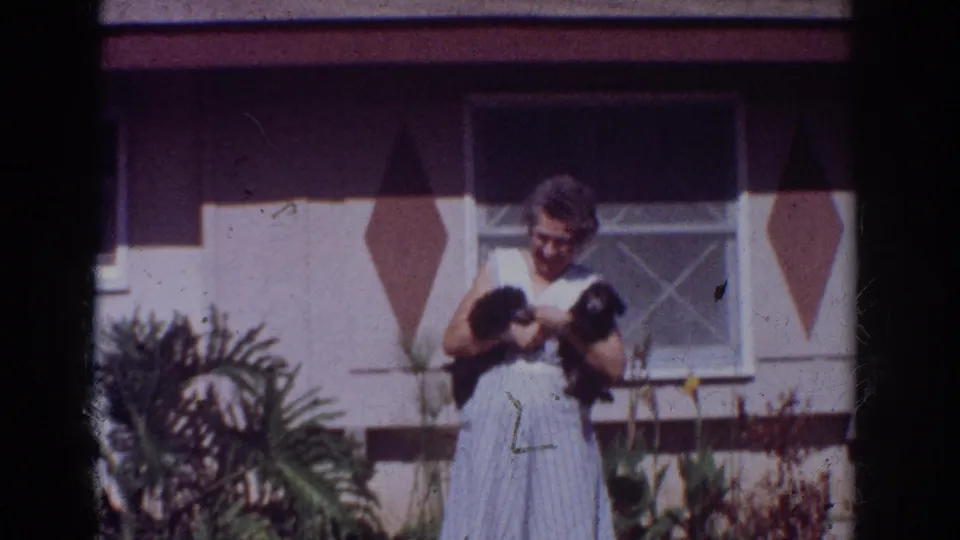 A woman smiles while holding two puppies in front of a patterned house exterior in the 1962 Yorba Linda home movie.