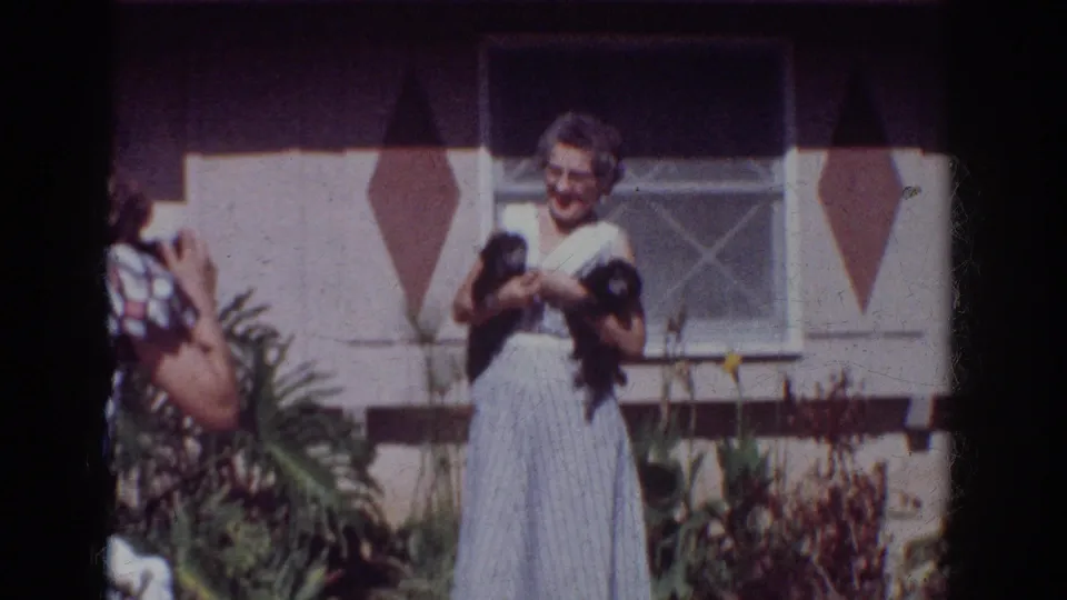 In this Yorba Linda 1962 home-movie frame, a second person with a camera appears as the woman poses with two small dogs outside the house.