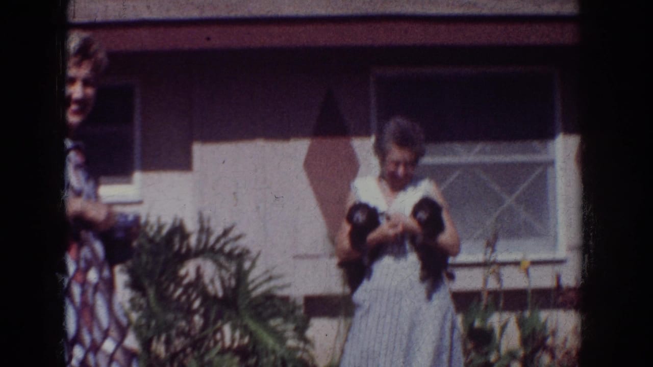 Older woman holding two small black puppies outside a house while another person stands partly at frame edge.