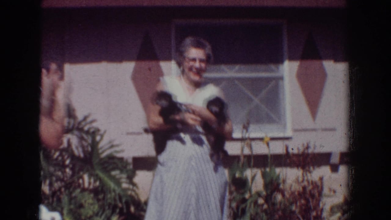 Smiling older woman holding two small black puppies in front of a house window and garden.