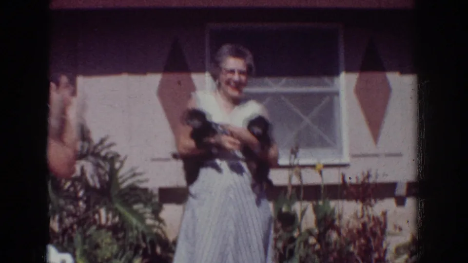 1962 Yorba Linda frame with an older woman smiling while holding two small black puppies outside a residential house.