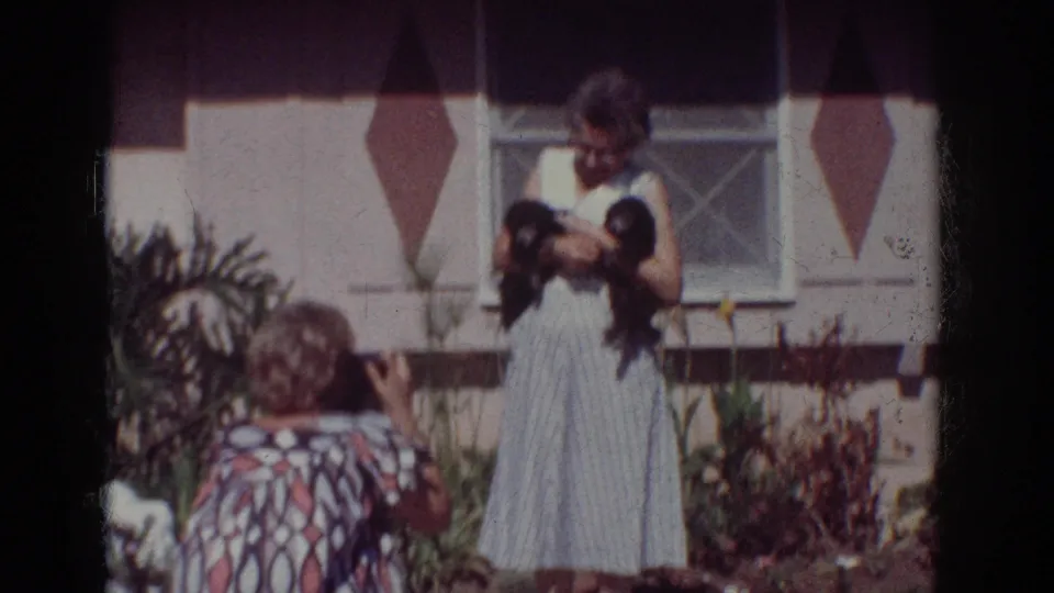 1962 Yorba Linda home-movie frame showing a kneeling woman taking a photo of an older woman holding two puppies.
