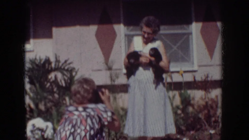 1962 Yorba Linda frame of a kneeling woman photographing an older woman holding two small black puppies in a residential garden.