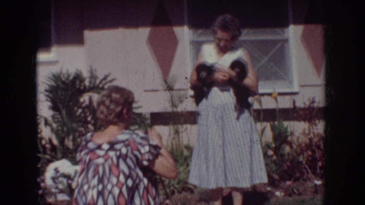 In a 1962 Yorba Linda home-movie frame, a woman holds two small black puppies while another woman photographs the scene in a garden.