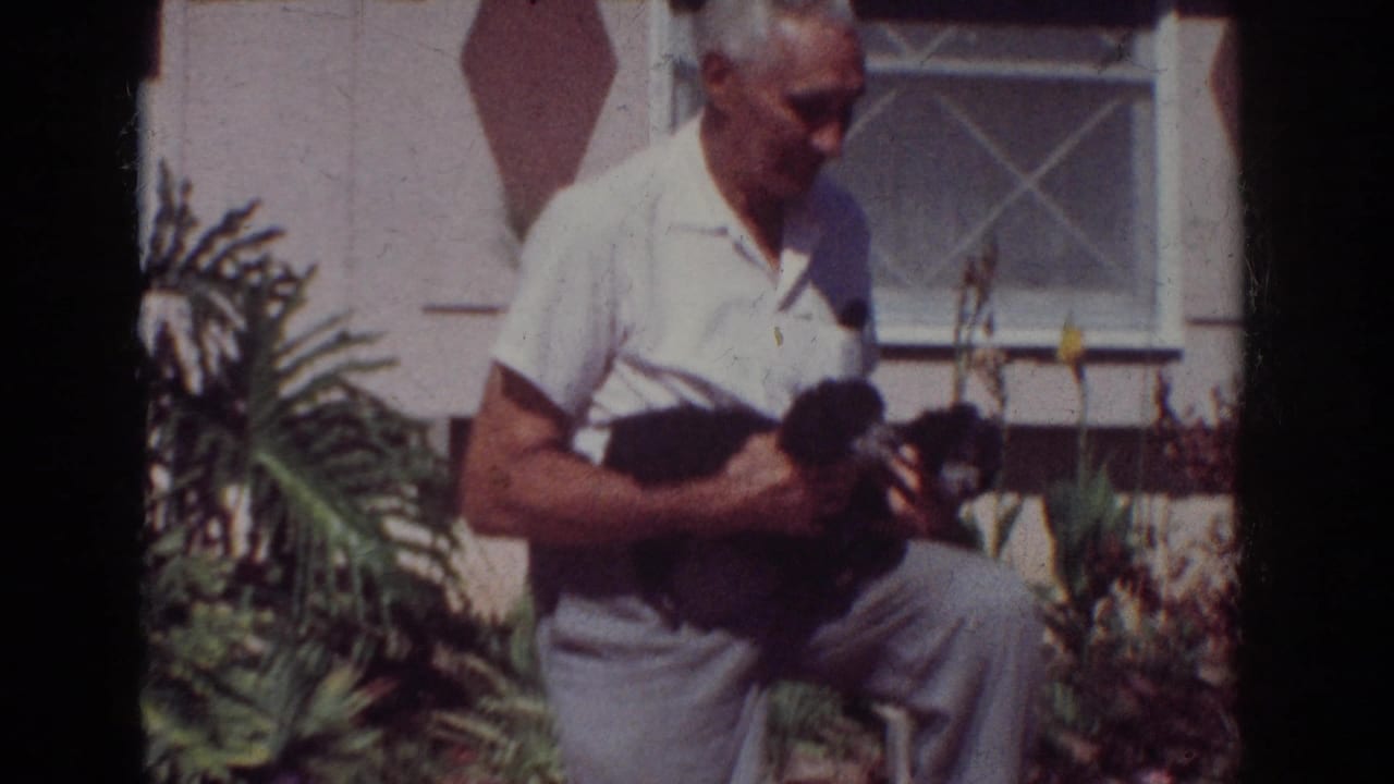 Older man kneels outdoors while holding two small black puppies near a house.