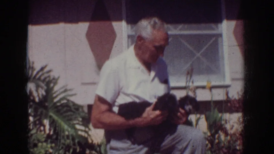 The older man continues holding the two puppies in the residential garden, with 8mm grain and soft color fading visible.