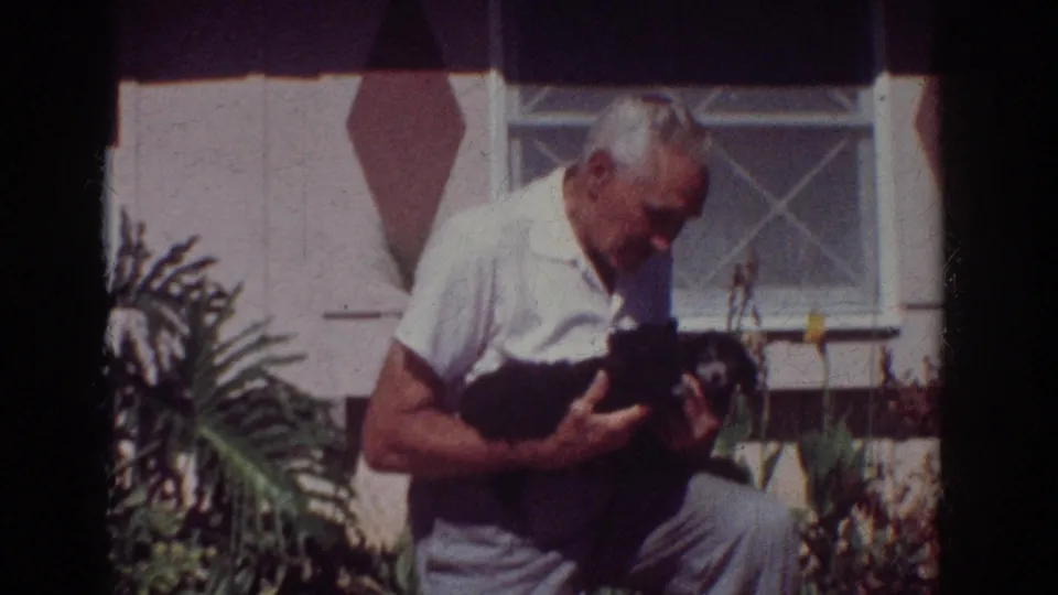 An older man cradles two small black puppies outside a Yorba Linda residence, framed by plants and a window in 1962 8mm footage.