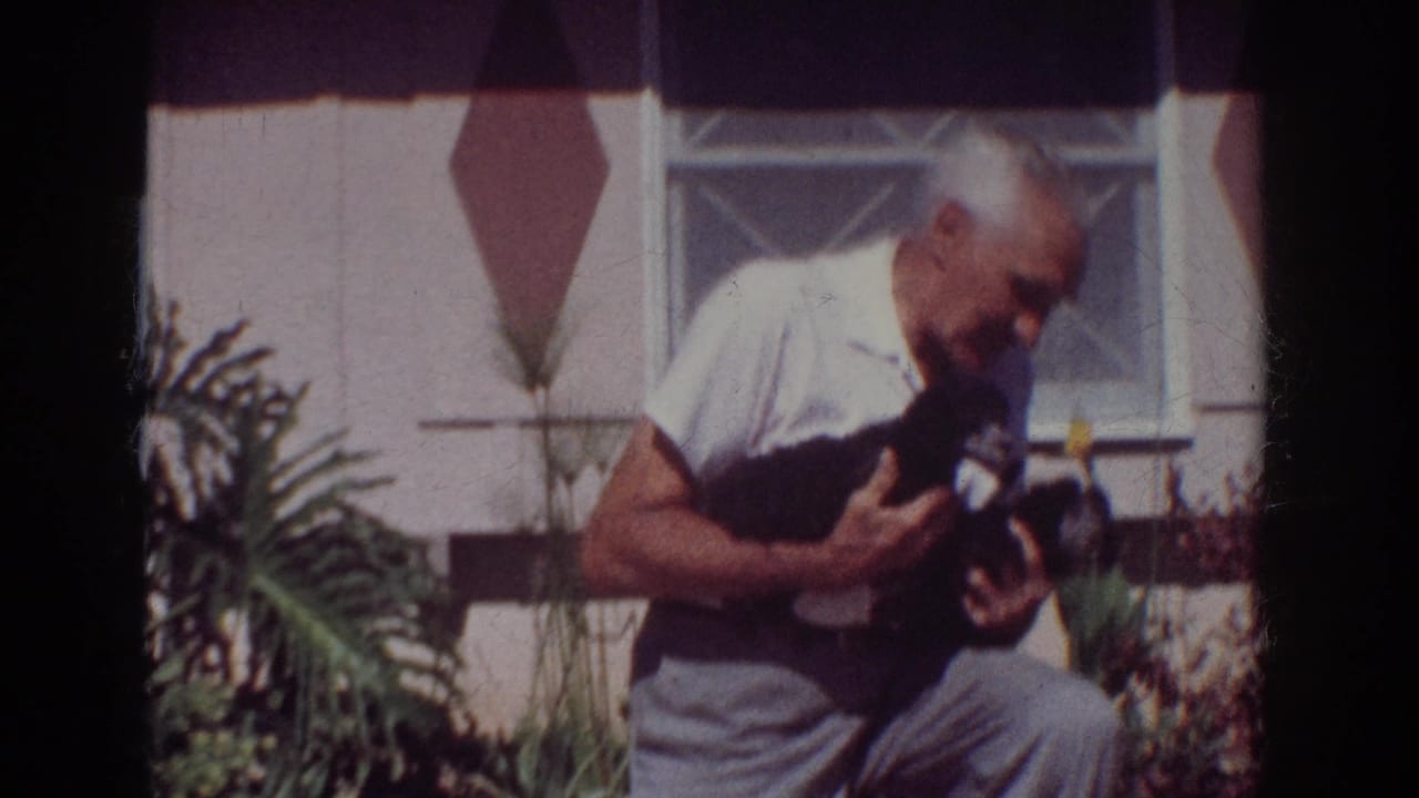 In supplied 1962 Yorba Linda 8mm footage, a man in a white short-sleeved shirt holds a small black puppy outside a suburban home.