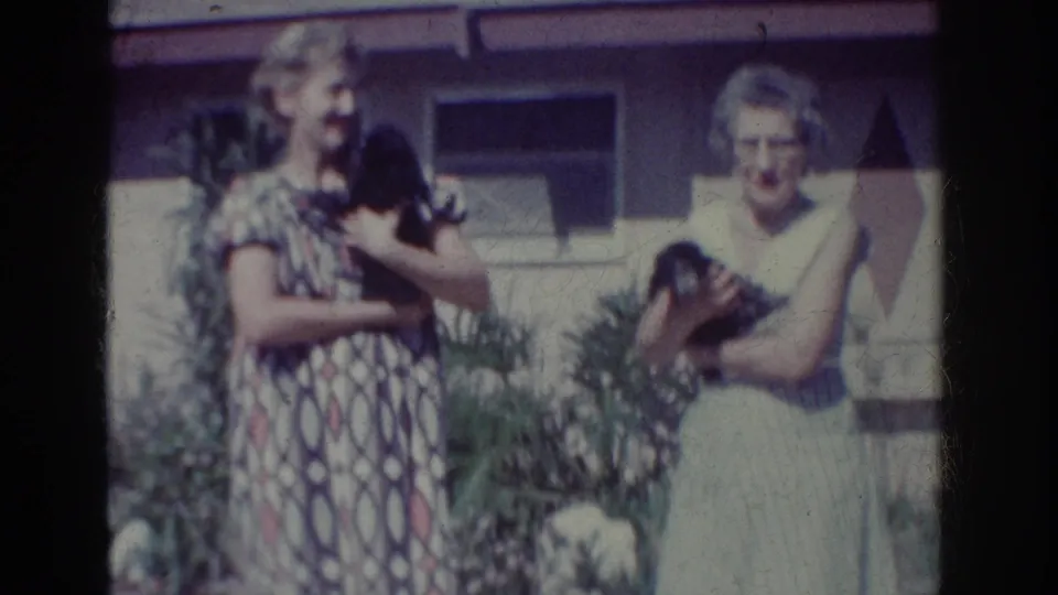Two women in 1960s dresses hold small black puppies in a Yorba Linda garden, with house windows and plants behind them.