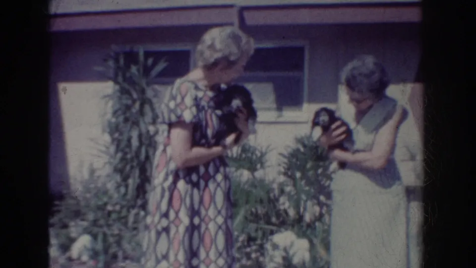 Two women in dresses look down at small black puppies in a suburban garden, framed by 8mm contact-sheet borders.