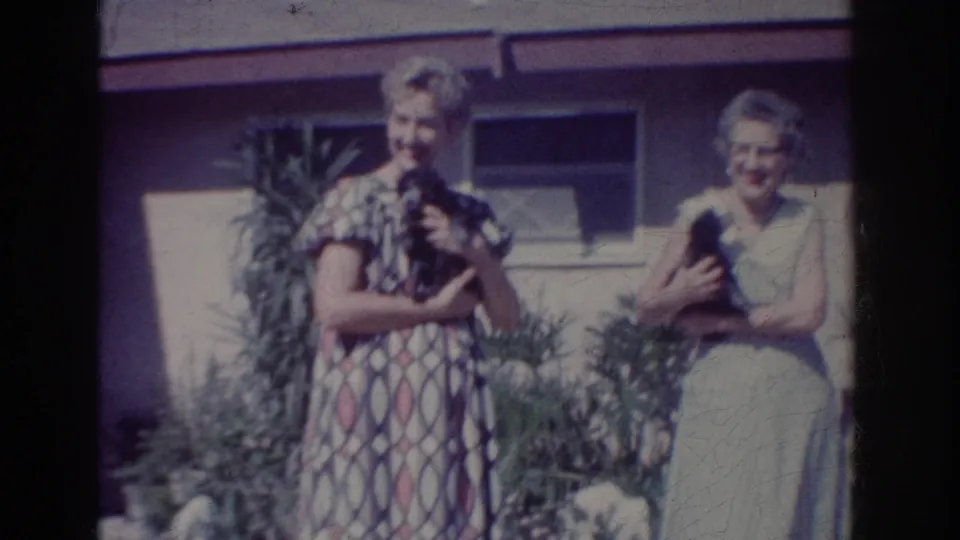 A 1962 home-movie frame shows two women smiling with puppies in a Yorba Linda residential yard.