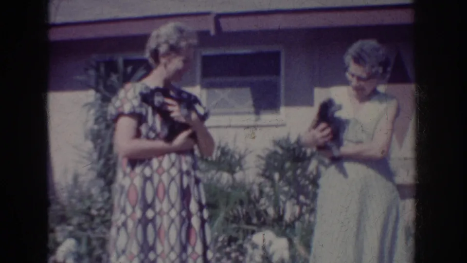 Two women with puppies stand in a Yorba Linda yard, one turning toward the other beside garden plants and a house exterior.