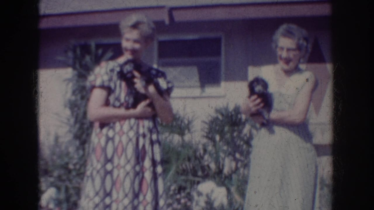 1962 Yorba Linda home-movie frame showing two women outside a house, each holding a small dark dog near garden plants.