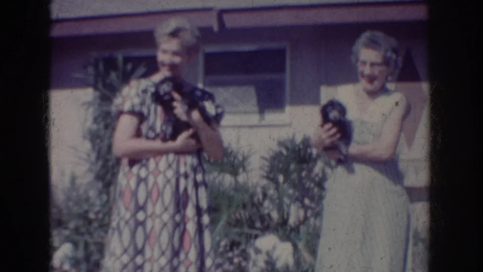 1962 Yorba Linda home-movie frame showing two women outside a house, each holding a small dark dog near garden plants.
