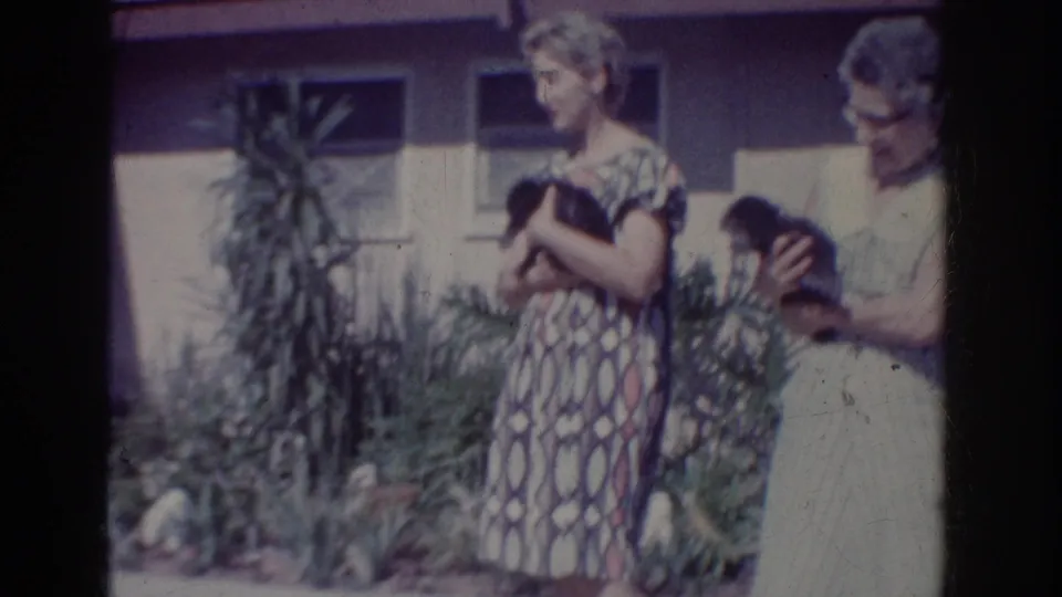 1962 Yorba Linda frame with two women holding small dark dogs beside planted landscaping and a house exterior.