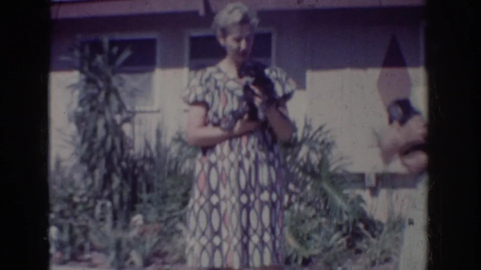 1962 Yorba Linda home-movie frame showing a woman in a geometric dress holding a small dog near garden plants and a house wall.