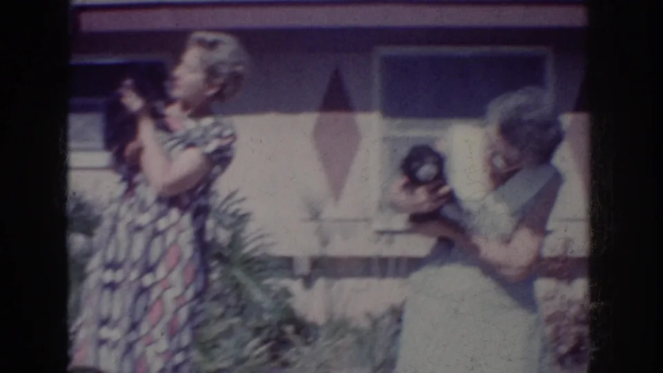 1962 Yorba Linda frame showing one woman lifting a small dark dog as another woman crouches nearby holding another dog.