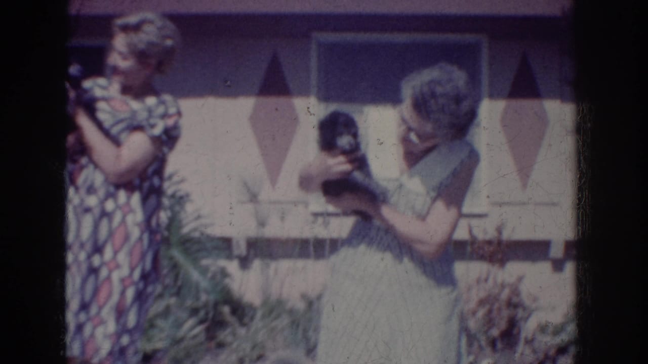 In 1962 Yorba Linda home-movie footage, two adult women stand in a yard holding small black dogs by a house facade.