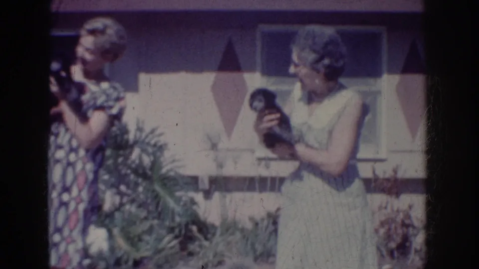 Two adult women in dresses hold small dogs outdoors in Yorba Linda, California, with a mid-century house behind them.