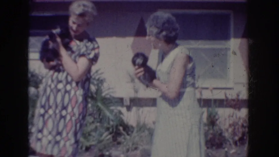 A woman in a geometric print dress cradles a small dog while another woman turns toward her in a 1962 suburban yard scene.