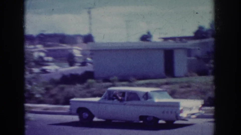 A white 1960s American sedan drives on a suburban Yorba Linda street with low buildings and parked cars in the background.