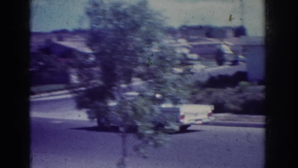 1962 Yorba Linda home-movie frame with a white car moving along a quiet residential street past trees and low houses.