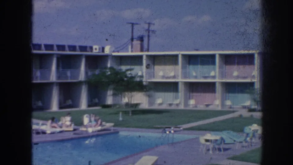 Mid-century Memphis motel exterior with pool, balconies, lawn, patio furniture, and guests relaxing poolside.