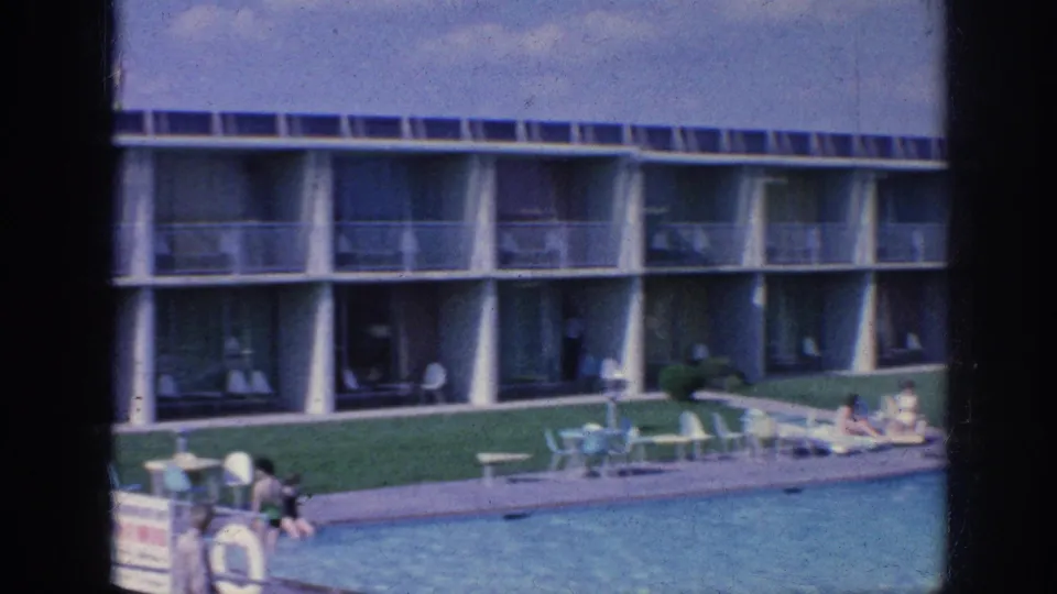 Memphis motel pool courtyard with rows of balconies, patio chairs, blue pool water, and several guests visible at a distance.