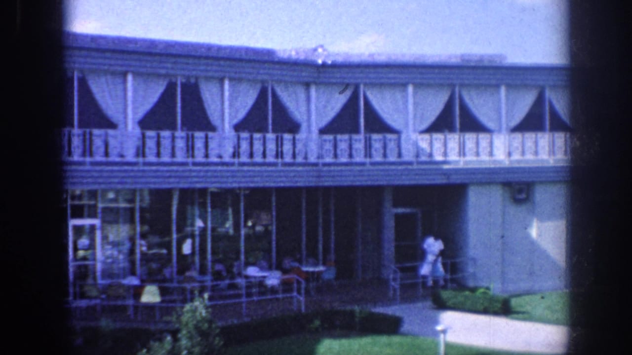 Two-story hotel exterior with balcony, porch seating, glass doors, and a man in white near the walkway.