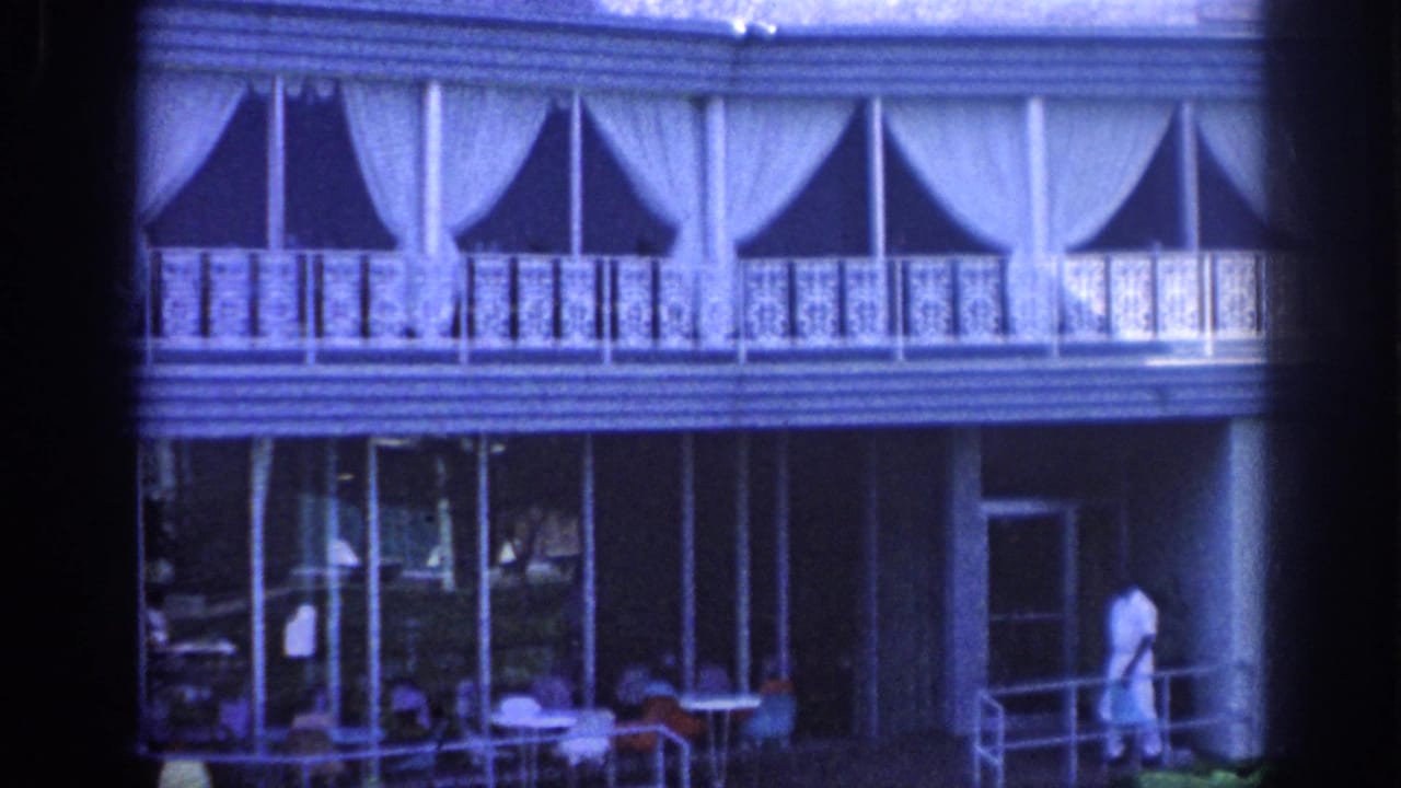 Hotel balcony and porch with draped upper windows, glass lower doors, patio furniture, and a man at right.