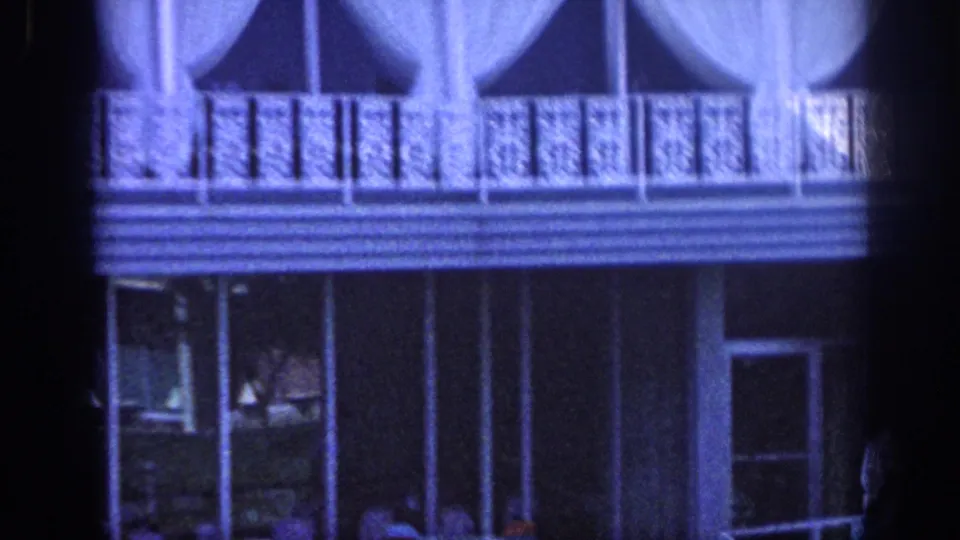 Closer 1963 Memphis hotel view emphasizing patterned balcony railings, white curtains, shaded glass doors, porch seating, and a partially visible figure at the edge.